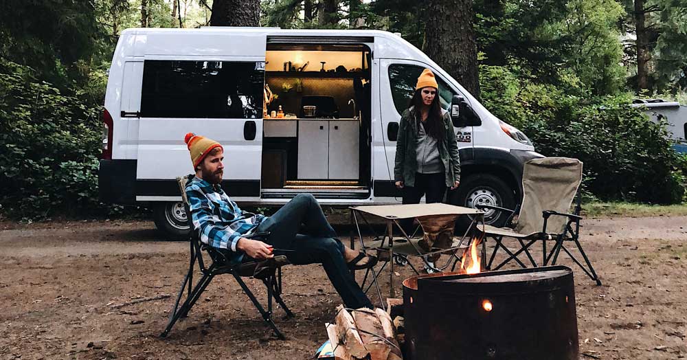A couple outside their campervan by a fire.