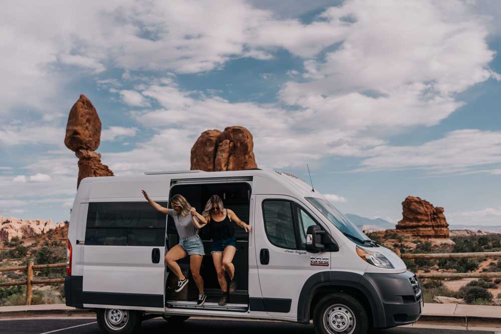 A couple jumping out of their campervan with desert rocks in the background.